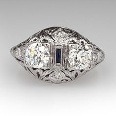 This Toi et Moi, circa 1920s, diamond ring features a pierced design with engraved details and is accented with two (2) old European cut diamonds set into four-prong settings, four (4), bead set, round single cut diamonds and one (1), bezel set, rectangular French cut synthetic blue sapphire. The ring measures 12.5mm at the top, rises 7.5mm above the finger, tapering to 1.7mm wide and 1.2mm thick at the base of the shank. It is currently a size 8. Antique Three Stone Jewelry For Formal Occasions, Antique Three Stone Diamond Ring For Formal Occasions, Art Deco Platinum Diamond Ring In Silver, Vintage Three Stone Platinum Jewelry, Blue Platinum Art Deco Diamond Ring, Formal Art Deco Three-stone Diamond Ring, Art Deco Three Stone Platinum Jewelry, Antique Platinum Three Stone Diamond Ring, Luxury Art Deco Sapphire Ring In Platinum