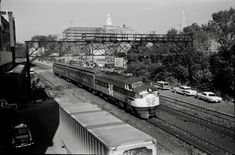 black and white photograph of train on tracks with cars parked along side it in front of large building