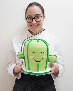 a woman holding up a cake with a green cactus on it's face and smiling