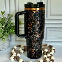 a black and gold travel mug surrounded by white and brown beads on a table next to a potted plant