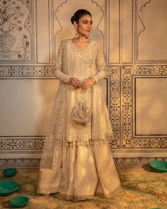 a woman standing in front of a wall wearing a white dress with gold embroidery on it