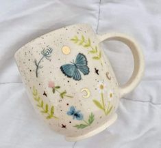 a ceramic mug with butterflies and flowers painted on it's side, sitting on a white sheet