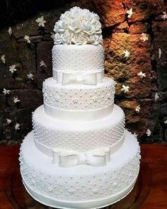 a three tiered wedding cake with white flowers and bows on the top is sitting on a wooden table