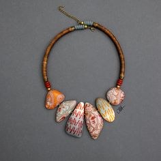 "Meet the chieftain" is a tribal inspired necklace. Warm, muted colors, like natural shades of browns and oranges are combined with reds and blue-grays. Abstract patterns and color gradients were used to decorate 6 different shaped beads that are placed together in a bib style necklace and form the front of the piece. 2 long cylindrical beads, wrapped in brown waxed cotton cord to enhance the natural look of the necklace, form the sides of the jewel.   "Meet the chieftain" is an everyday, friend Handmade Unique Orange Necklaces, Unique Handmade Orange Necklaces, Unique Orange Necklace With Wooden Beads, Multicolor Bohemian Beaded Necklace With Boho Collar, Bohemian Multicolor Beaded Necklace With Boho Collar, Earthy Red Handmade Jewelry, Orange Bohemian Beaded Necklaces For Jewelry Making, Bohemian Orange Beaded Necklaces For Jewelry Making, Orange Bohemian Beaded Necklaces