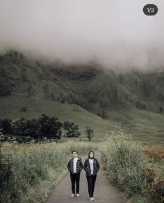 two people walking down a path in the middle of a field with mountains behind them