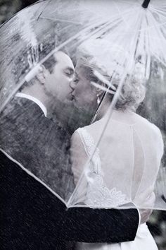 a bride and groom kissing under an umbrella on their wedding day, in the rain