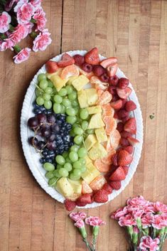 a platter filled with lots of fruit on top of a wooden table next to pink flowers