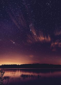 the night sky is filled with stars and clouds over a lake in front of a forest