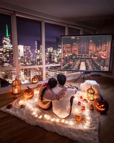 two people sitting on a rug in front of a large window with city lights and pumpkins