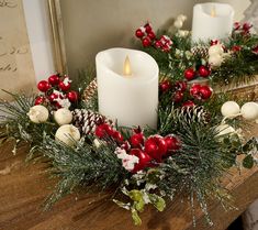 two white candles sitting on top of a wooden table covered in greenery and red berries