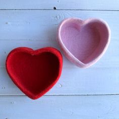 two felt heart shaped bowls sitting next to each other on a white wooden surface, one red and the other pink