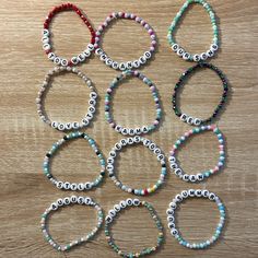 six bracelets with beads and letters on them sitting on a wooden surface, all in different colors