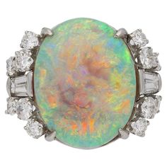 Vintage opal and diamond ring. Set with one oval cabochon natural unenhanced white opal in an open back four claw setting with an approximate weight of 10.00 carats, flanked by twelve round brilliant cut diamonds in open back claw settings with a combined weight of 1.00 carats, further flanked by two tapered baguette cut diamonds in open back half rubover settings with a combined weight of 0.60 carats. The total approximate diamond weight is 1.60 carats, to an impressive solitaire design with a Unique Opal Ring, Opal And Diamond Ring, Opal Ring Vintage, Australian Black Opal, Vintage Cocktail Ring, Diamond Cocktail Rings, Gemstone Jewellery, Exclusive Jewelry, Baguette Cut