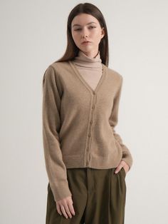 a woman standing in front of a white wall wearing a brown sweater and khaki pants