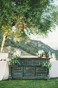 an old dresser is decorated with greenery and wine bottles for a rustic outdoor bar