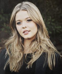 a woman with long blonde hair standing in front of some trees and looking at the camera