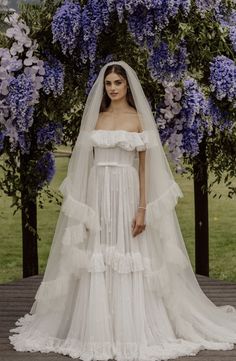 a woman in a white wedding dress standing on a wooden platform with purple flowers behind her