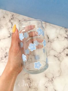 a hand holding a glass with blue flowers on it and yellow nail polish in front of a marble counter top