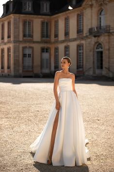 a woman standing in front of a large building wearing a white wedding dress with high slit