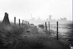 a black and white photo of a foggy field with industrial buildings in the background