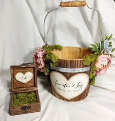 a wooden basket with moss and flowers in it next to a ring box on a white sheet