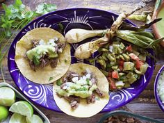 three tacos on a blue plate with limes and other food items around it