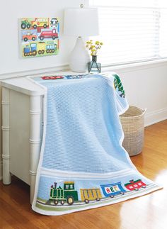 a blue crocheted baby blanket with trains on it sitting next to a lamp