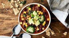 a white bowl filled with chili, corn and avocado on top of a wooden table