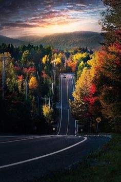 an empty road surrounded by trees and hills in the background at sunset or sunrise with clouds