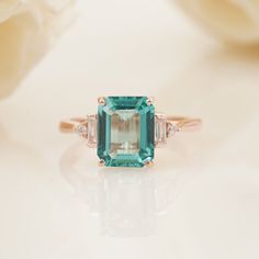 an emerald and diamond ring sitting on top of a white table with flowers in the background