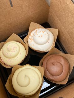 four different colored cupcakes in brown boxes on a black tray with white frosting and sprinkles