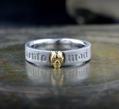 This gold ring is a narrow, elegant band that combines symbolism and style. Its minimalist design belies its deep meaning, capturing attention with an intriguing central motif: a finely sculpted skull that sits precisely on the surface of the ring. The skull, rendered in detail, evokes a sense of mystery and a constant reminder of mortality. On the outside of the band, the phrase "Memento Mori" is engraved in careful, well-defined letters, reminding the wearer of the impermanence of life. This inscription, meaning "Remember that you will die," serves as a philosophical reflection and a call to live with purpose and awareness. The shine of the gold adds a touch of luxury, while the simple design of the band ensures that the ring is comfortable to wear. This ring is ideal both as a meaningfu Silver Stackable Wedding Rings Tarnish Resistant, Symbolic Engraved White Gold Skull Ring, Classic Formal Skull Ring With Polished Finish, Classic Skull Ring As Gift With Polished Finish, Stainless Steel Jewelry With Engraving For Promise, Symbolic Engraved Stackable Rings For Anniversary, Adjustable Round Skull Ring For Anniversary, Symbolic Stainless Steel Engraved Ring For Anniversary, Adjustable Skull Ring For Anniversary