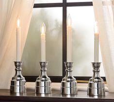 three silver candles sitting on top of a table next to a window with white curtains