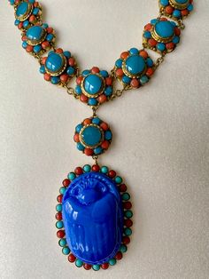 A vintage striking and beautiful  1930's Egyptian Revival coral and turquoise glass Scarab drop necklace. A single row of turquoise and coral glass cabochon rosettes, 15 in total, with a pendant of a further rosette and a large turquoise glass Scarab Beetle with a surround of turquoise and coral glass cabochons. The metal mounts are gilt metal and the cabochons collet set. A lovely clasp set with 3 glass turquoise cabochons, hook and ring clasp secures. Length of 17 inch with 4.1/2 inch drop. In Antique Blue Cabochon Necklace, Traditional Blue Cabochon Necklace, Traditional Blue Cabochon Necklaces, Egyptian Revival Jewelry, Czech Jewelry, Czech Glass Necklace, Exotic Jewelry, Scarab Beetle, Egyptian Jewelry