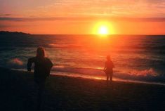 two people standing on the beach watching the sun go down over the ocean with waves