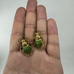 a pair of green and gold beetle earrings on a person's left hand in front of a white background