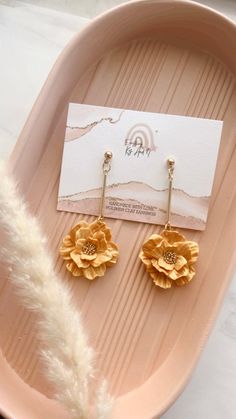 the gold flower earrings are next to a white feather on a pink platter with a business card