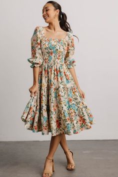 a woman in a floral print dress smiles while standing against a white wall with her hand on her hip