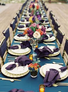 a long table set with place settings and purple napkins on it, along with flowers