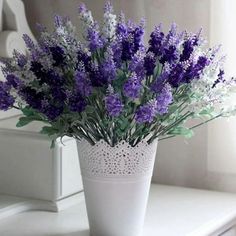 a white vase filled with purple flowers sitting on top of a table next to a window