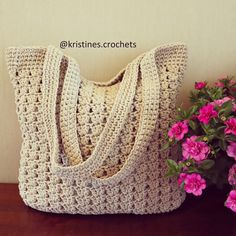 a crocheted purse sitting next to pink flowers on a wooden table in front of a white wall