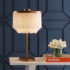 a lamp on a table next to a book and vase with white flowers in it