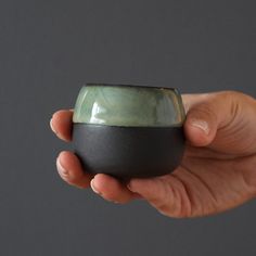 a hand holding a small green and black cup in it's left hand, against a gray background