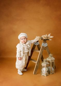 a baby standing next to a ladder with flowers on it's head and wearing a white outfit