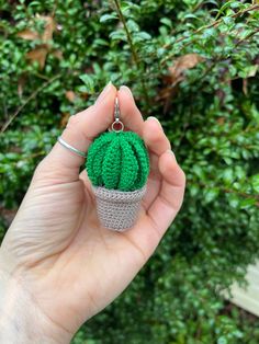 a hand holding a green crocheted cactus keychain in front of some bushes