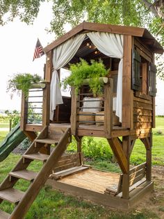 a wooden play house with a slide and green plants in the top floor window boxes