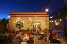 people are sitting at tables outside in front of a building with lights strung over it