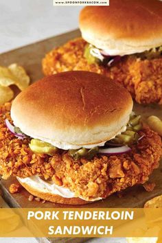 two fried chicken sandwiches sitting on top of a wooden cutting board next to potato chips