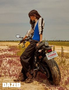 a woman standing next to a motorcycle in the middle of a field with purple flowers