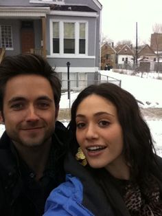 two people standing in front of a house with snow on the ground and houses behind them